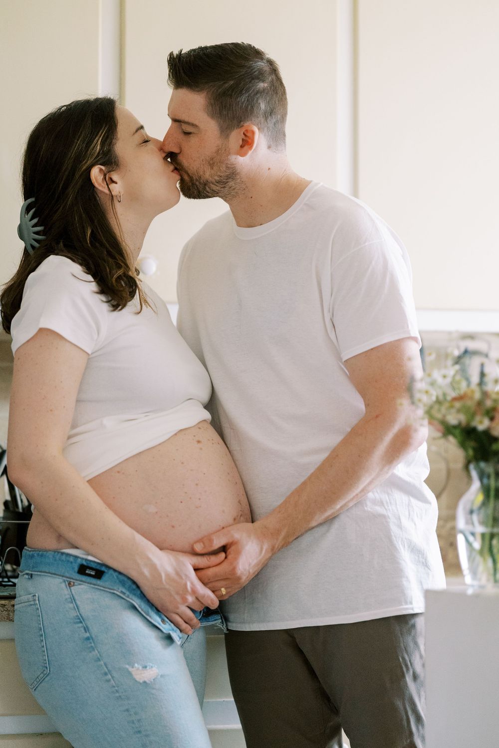 Intimate moment between expecting couple in white shirts and jeans embracing near window in Clifton, New Jersey.