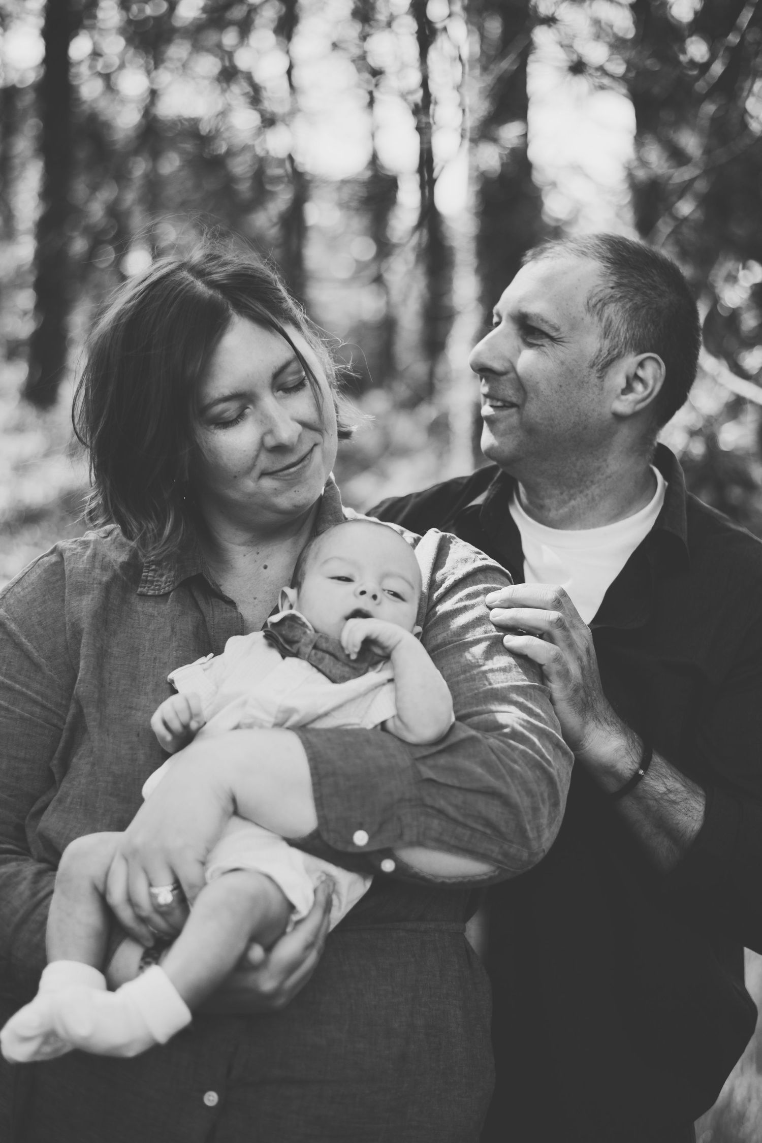 A sweet black and white family portrait taken outdoors in a forest with soft bokeh lighting effects in the background.