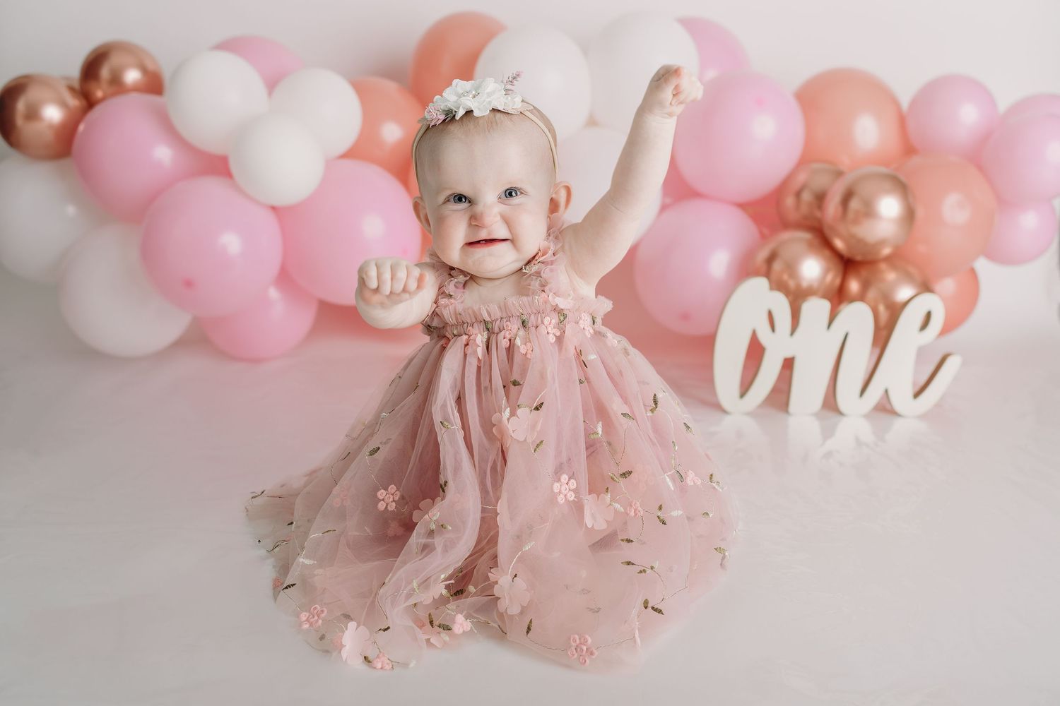 A baby in a pink tulle dress celebrates their first birthday with balloon decorations in soft pink, white and rose gold.