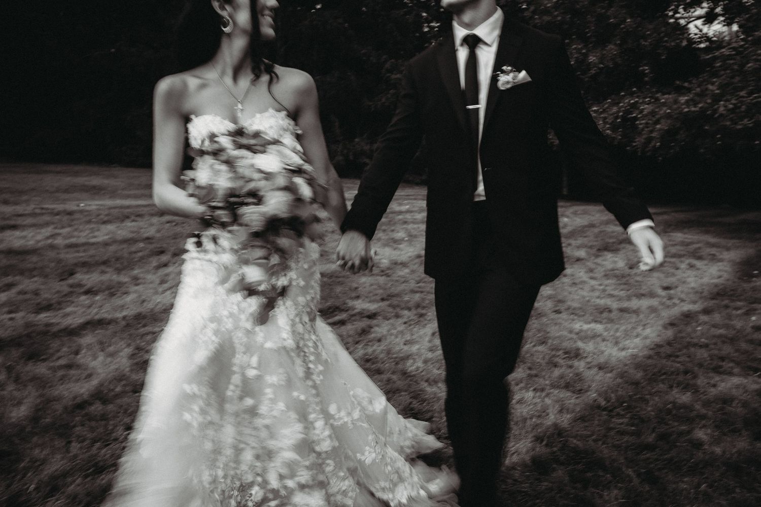 Black and white photo of a bride and groom walking together outdoors, with the bride holding a floral bouquet.