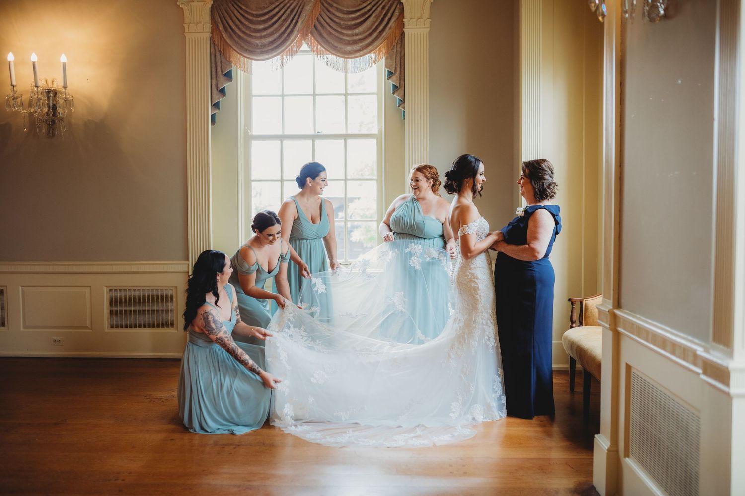 A group of bridesmaids in light blue dresses help adjust a long white veil in an elegant room with wall sconces.