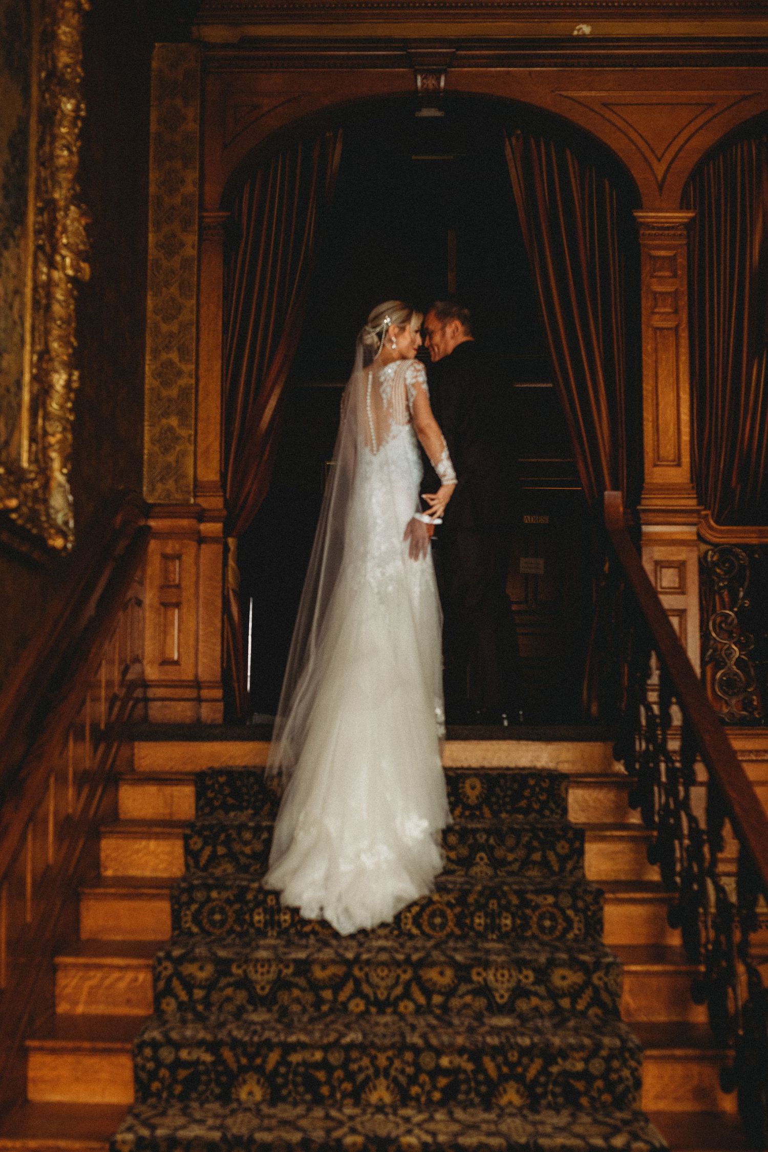 A wedding dress silhouette stands elegantly on an ornate staircase with dark carpeting and wooden trim in warm lighting.