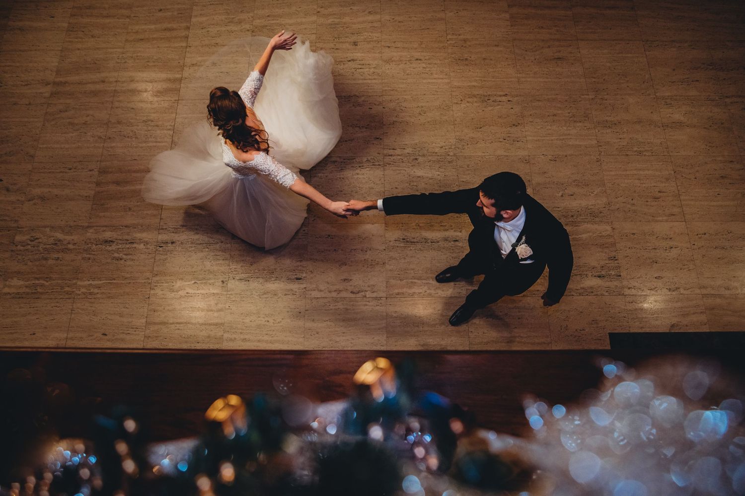 A bride and groom dance together on a wooden floor captured from above with dreamy bokeh lights in the foreground.