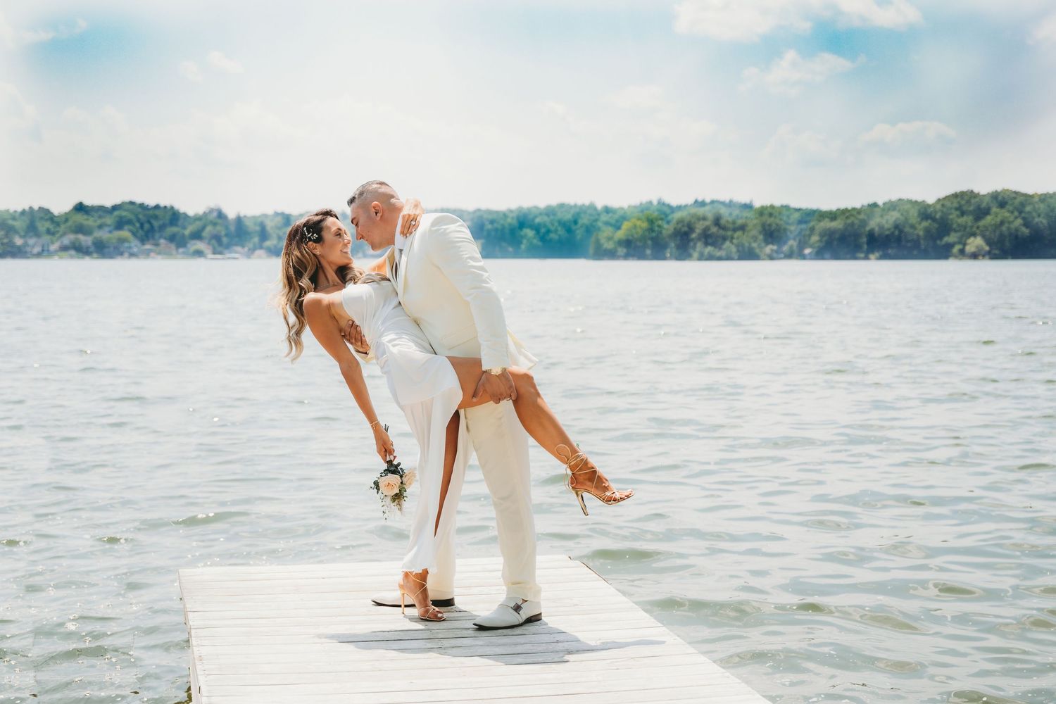 A romantic wedding photo on a dock by a lake with white attire and dramatic dip pose.