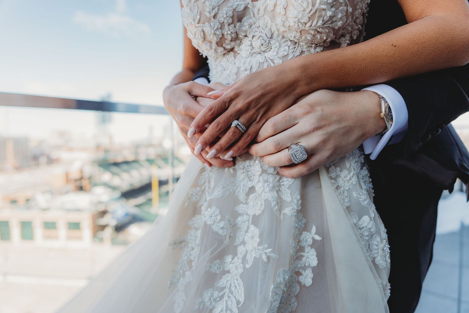 Close up of hands showing wedding ring detail with lace wedding dress in background.
