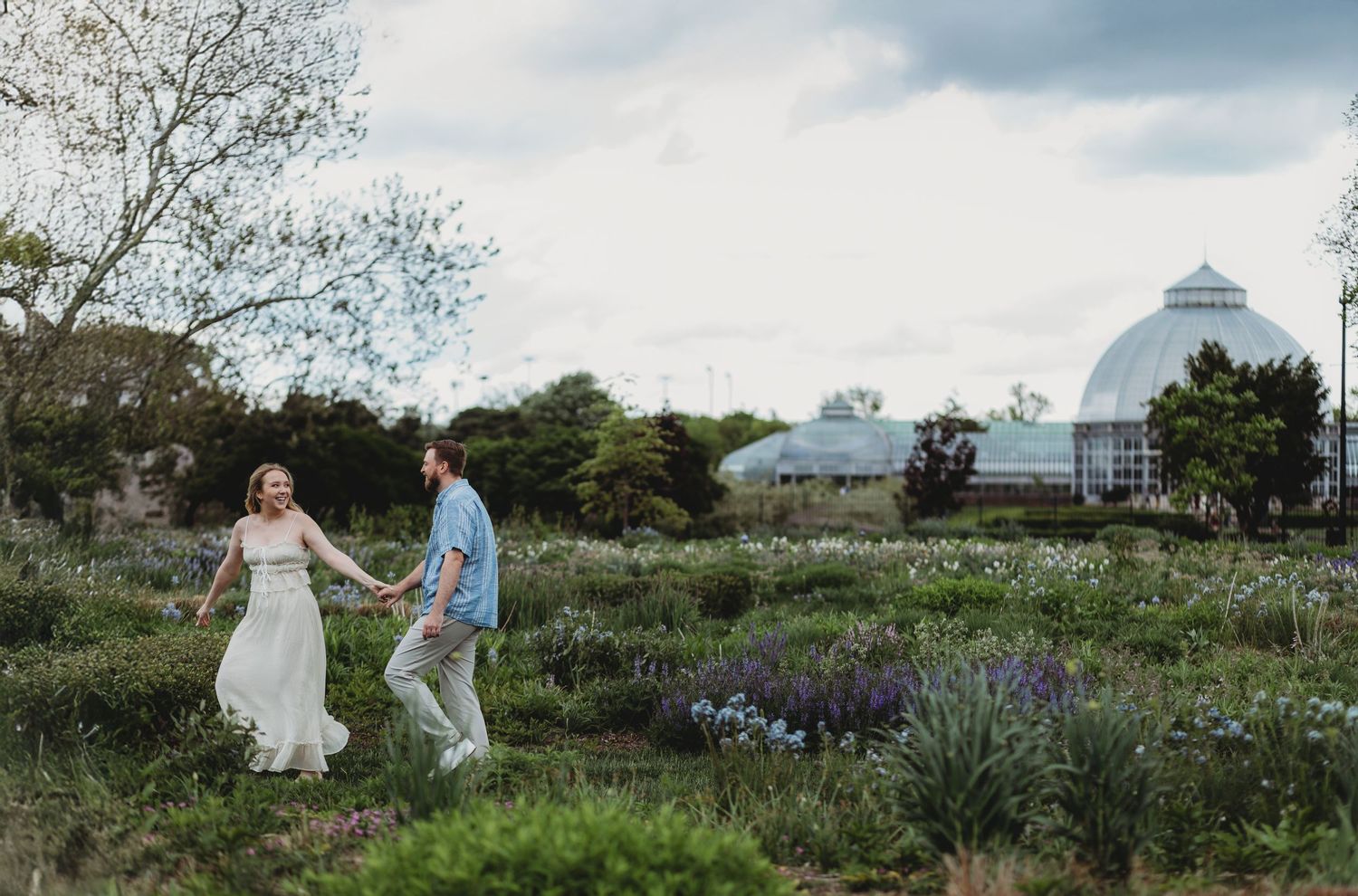Two people walk through a lush garden with purple wildflowers, with a conservatory dome visible in the background.