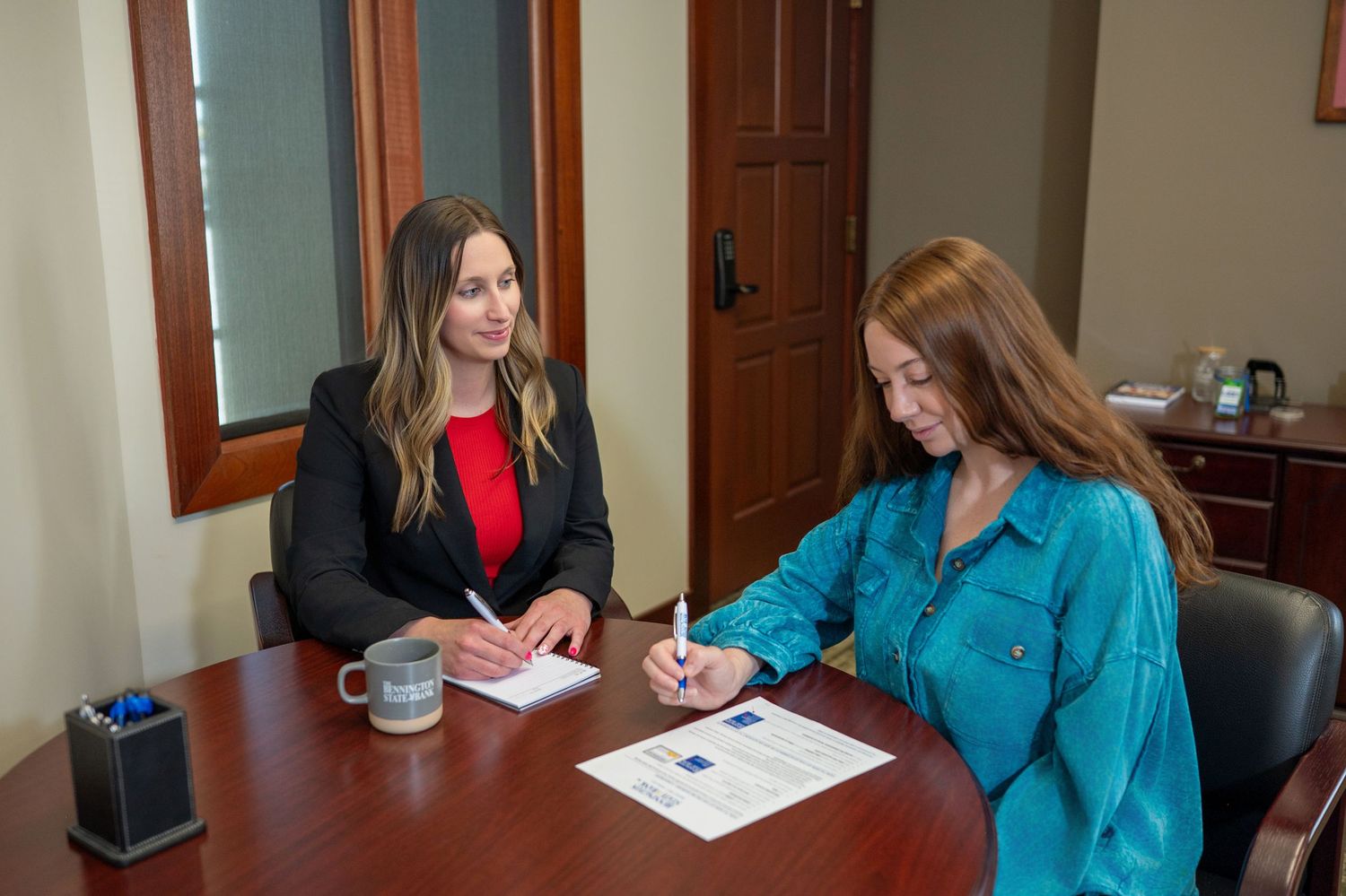 Two people sit at a desk in a professional office setting reviewing paperwork together over coffee.