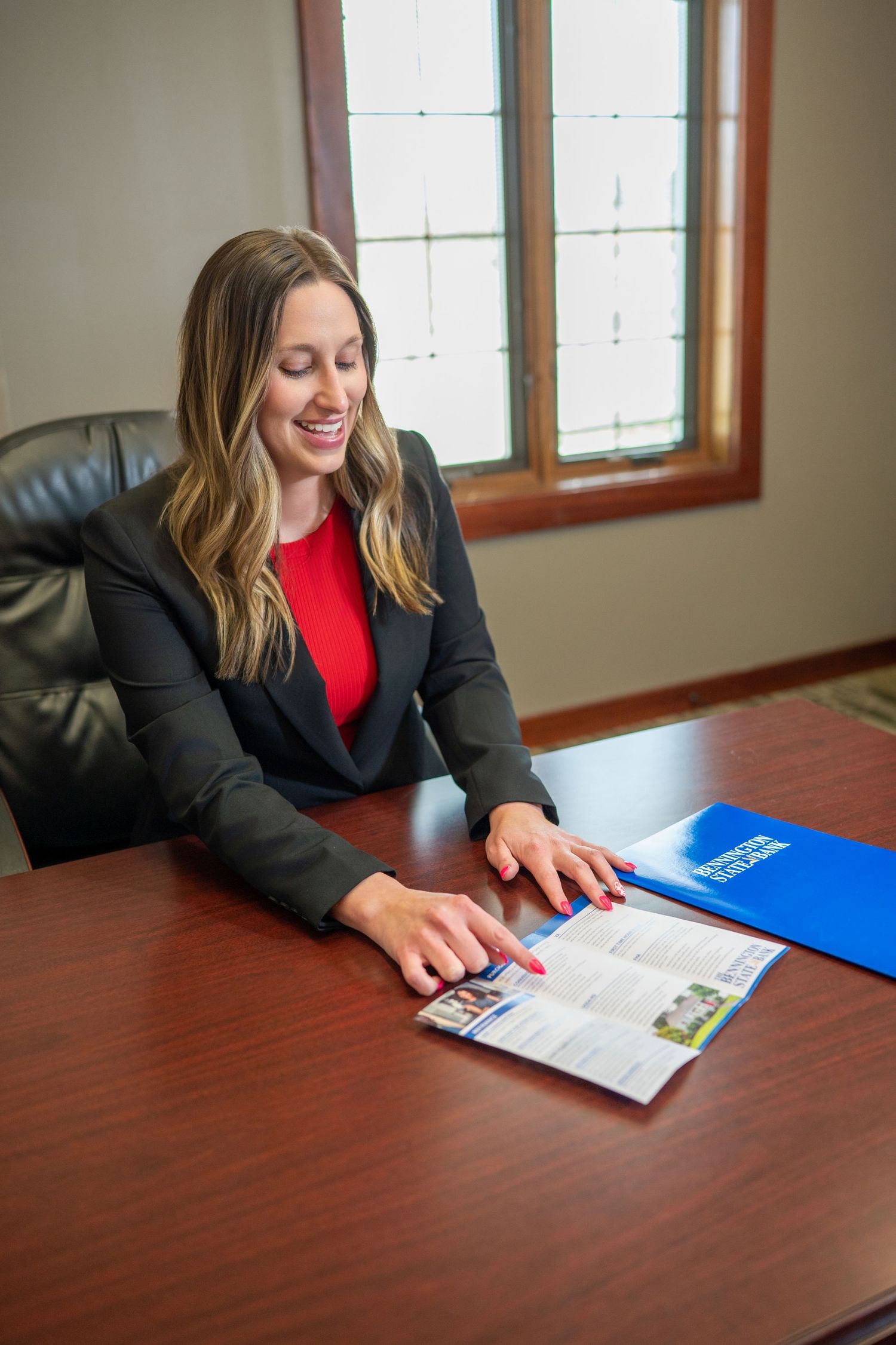 Professional in a dark blazer and red top sits at desk reviewing documents with a blue folder on wooden table.