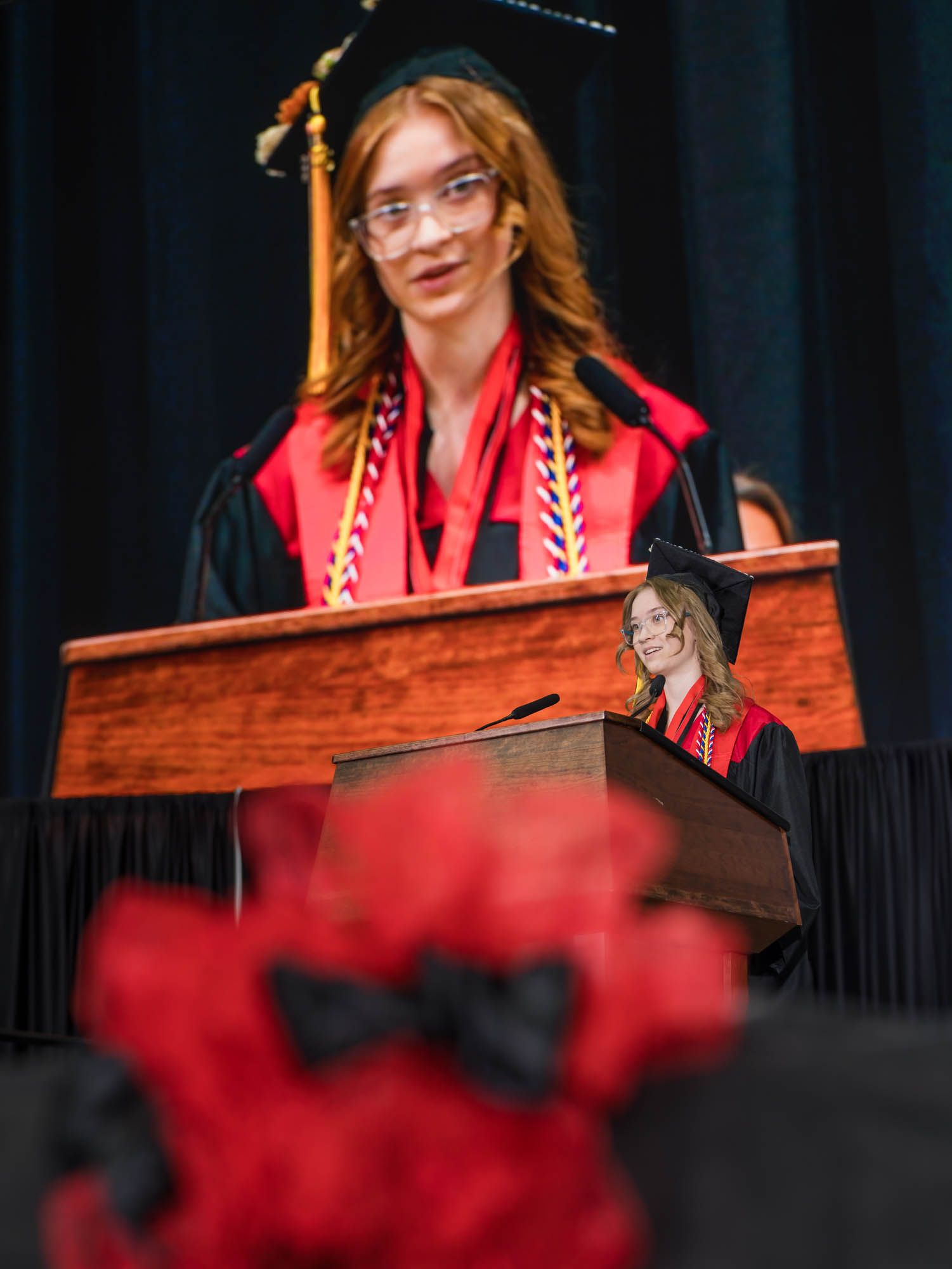 A graduate in academic regalia stands behind a wooden podium with honors cords and regalia during commencement ceremony.