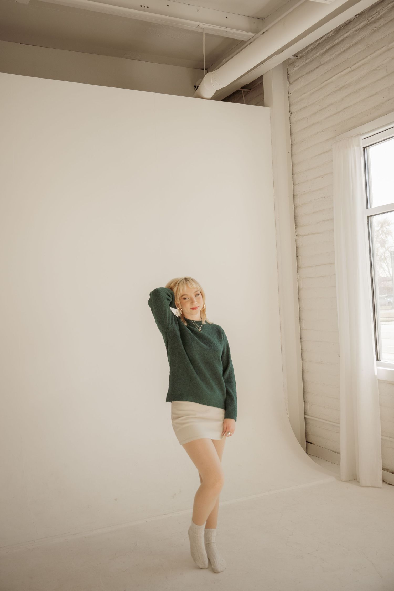 A person in a green sweater and beige shorts poses against a white wall in a bright minimalist studio setting.