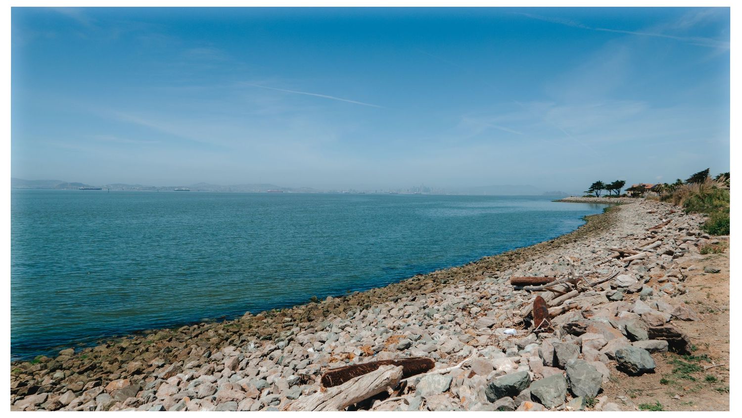 A rocky shoreline meets the tranquil blue waters of the ocean under a clear sky with distant cliffs in view.