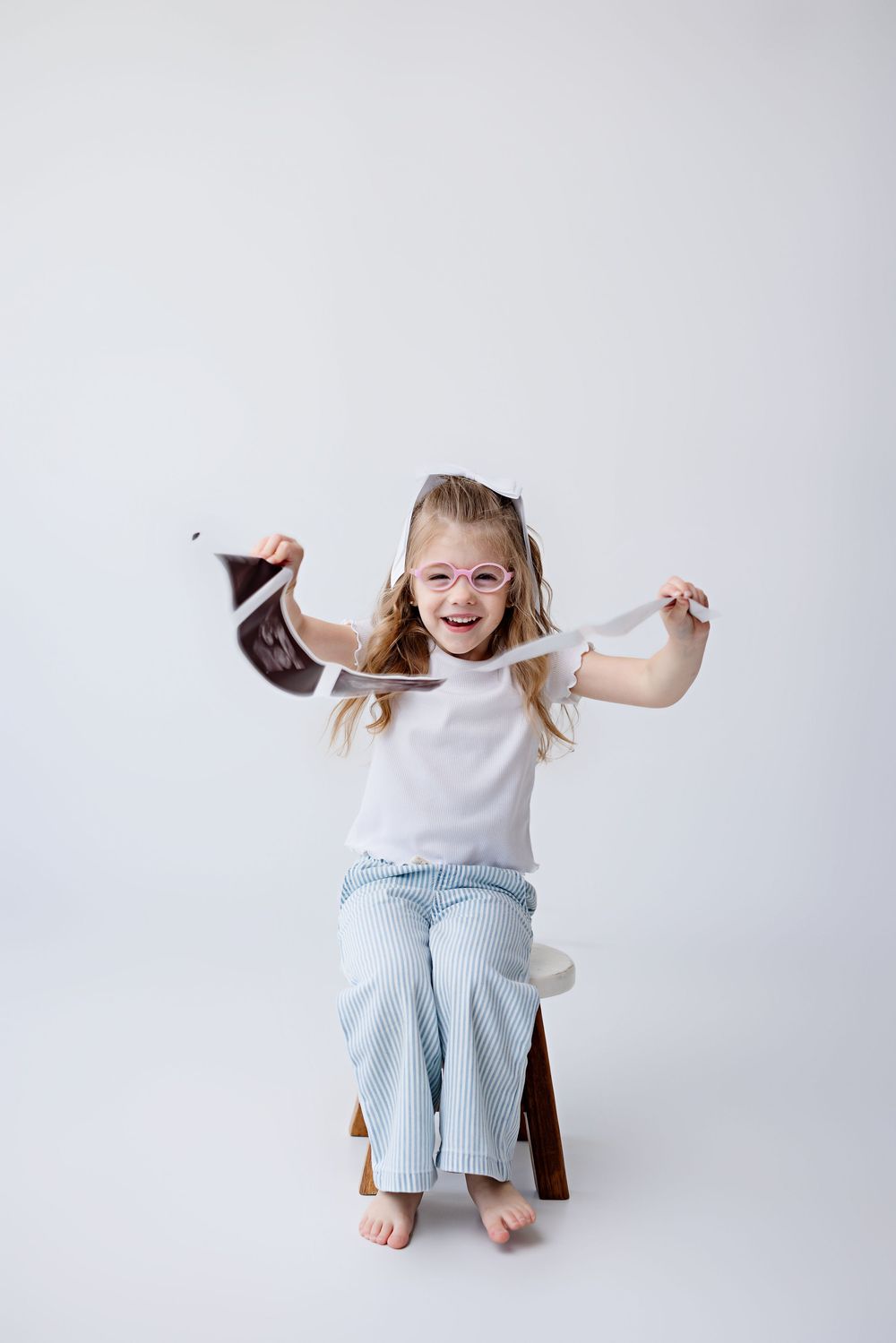 Joyful child in white tank top and light jeans sits on wooden stool against white background while playing with hair.
