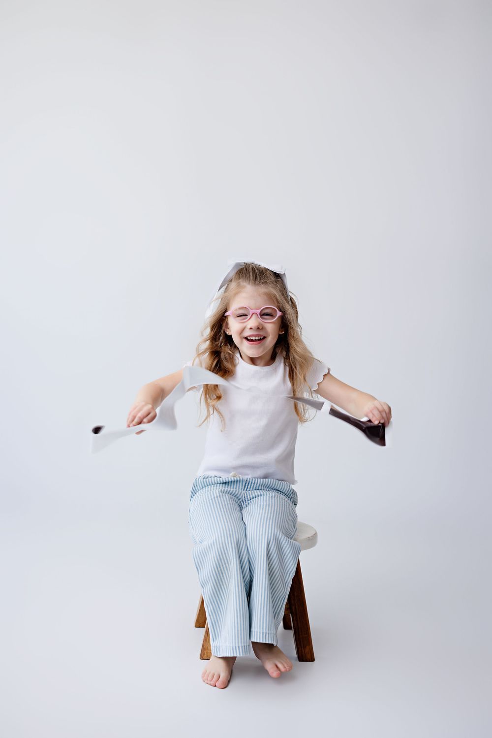 Energetic young person in white shirt and jeans sitting on wooden chair against light background, laughing joyfully.
