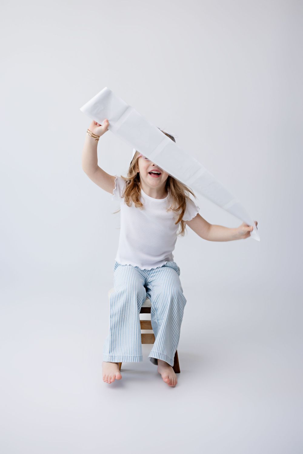 Playful child in light blue pants and white shirt holding up a large piece of white paper while sitting on wooden chair.