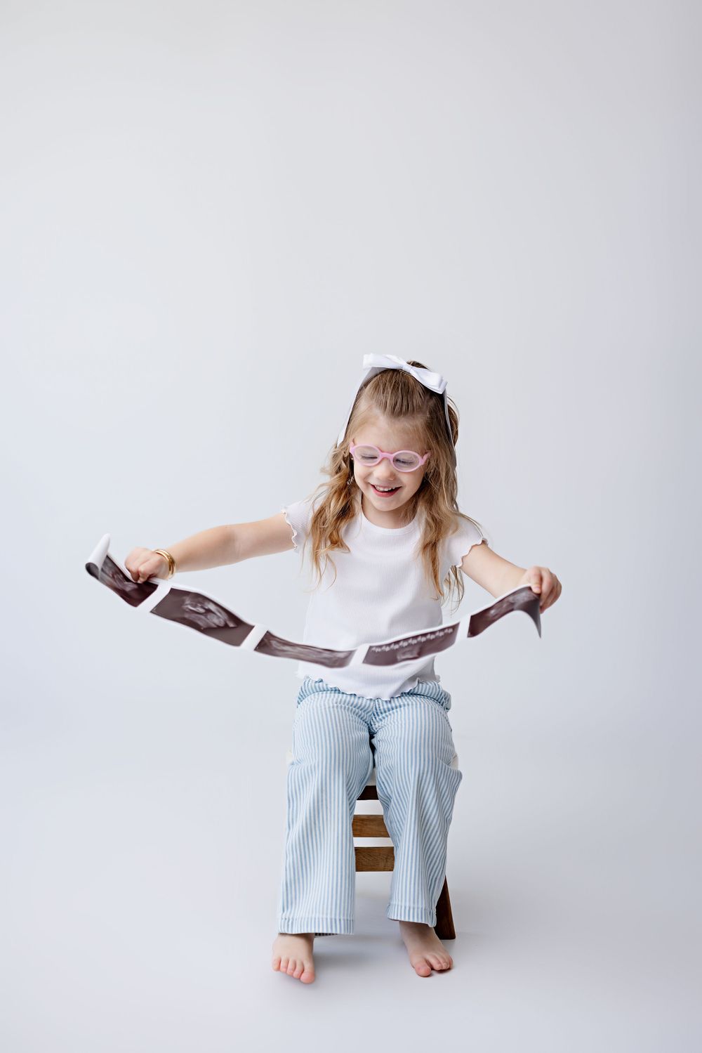 Child sitting on wooden box wearing white top and striped pants laughs while holding open a book against gray background.