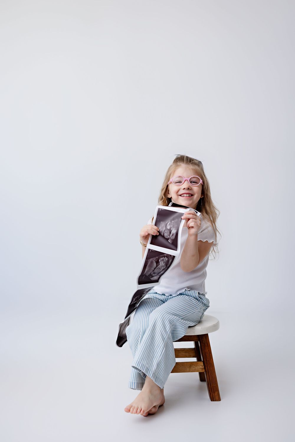 A joyful child in glasses sits on a wooden stool holding an ultrasound photo against a white background.