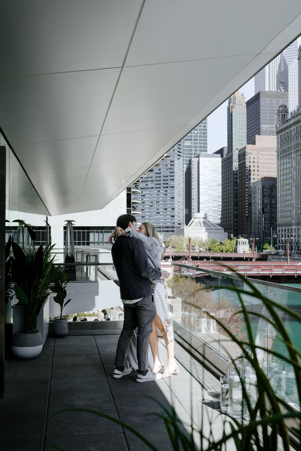 Modern couple embraces on urban balcony overlooking Chicago River with city skyline and skyscrapers in background.