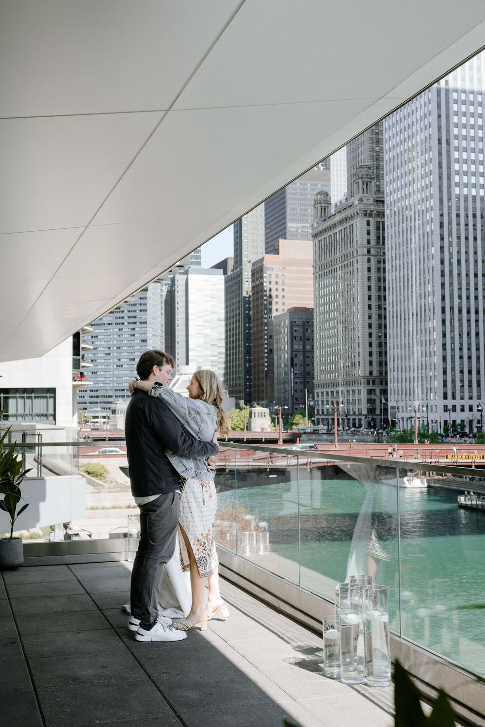 A couple embraces while overlooking the Chicago River with iconic skyscrapers and modern architecture in the background.
