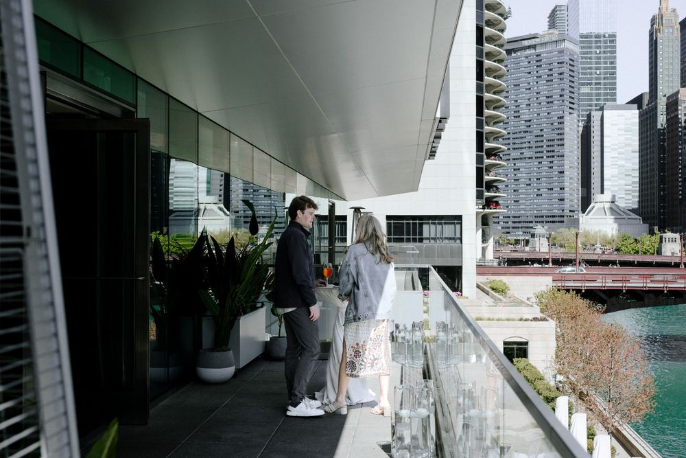People walking on a modern balcony terrace with glass railings overlooking an urban cityscape with high-rise buildings.