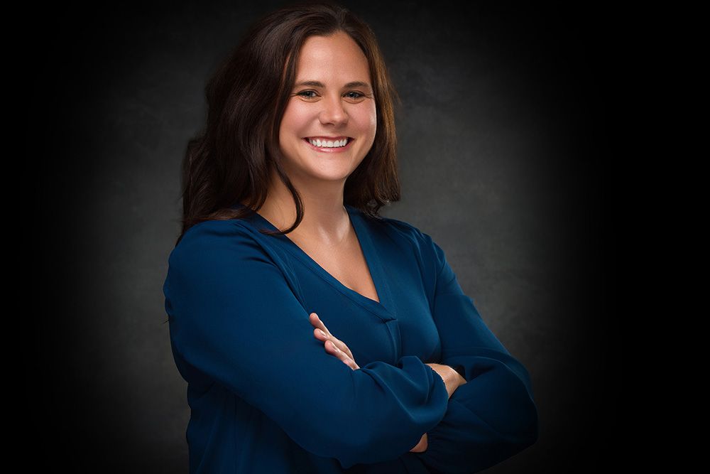 Professional business portrait of someone in a navy blue top with arms crossed against a dark studio background.