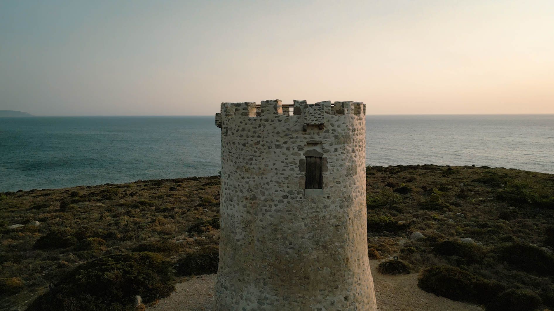 Ancient stone watchtower standing on a rugged coastal cliff overlooking the calm Mediterranean Sea at sunset.
