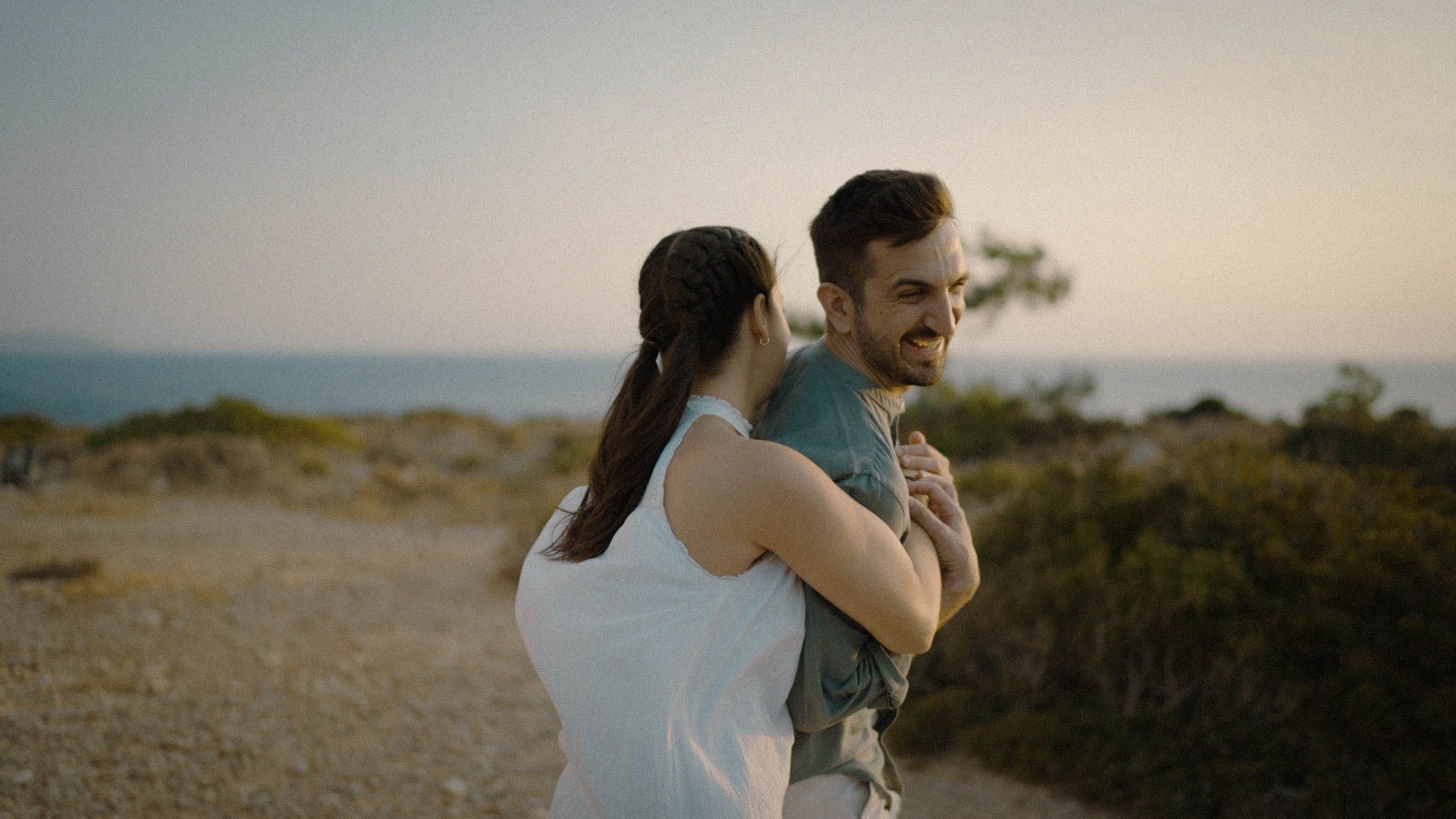 A couple embraces on a hilltop trail at sunset, surrounded by coastal brush and ocean views.