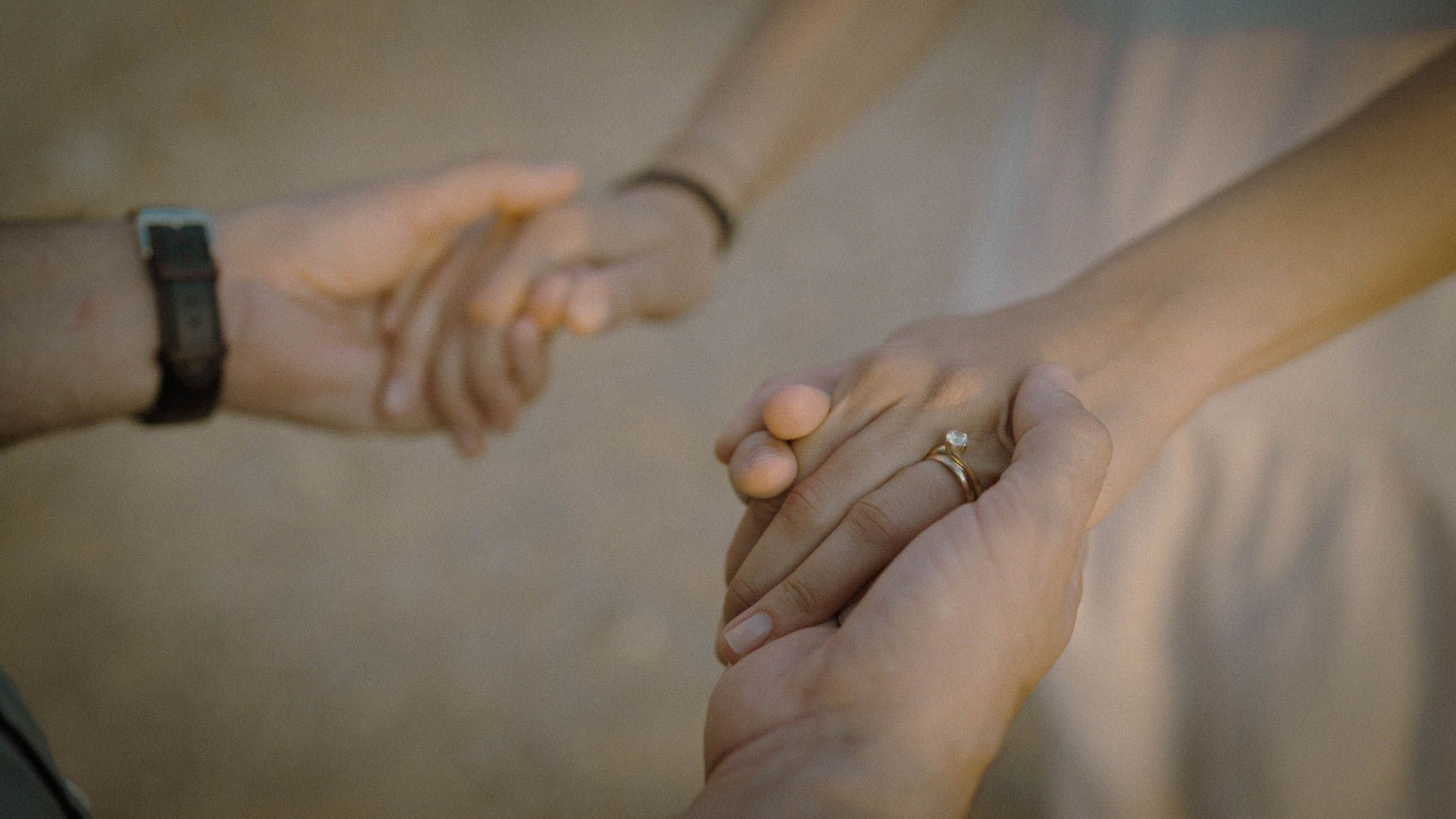 Two hands tenderly clasped together, one wearing a watch and the other adorned with a ring in soft lighting.