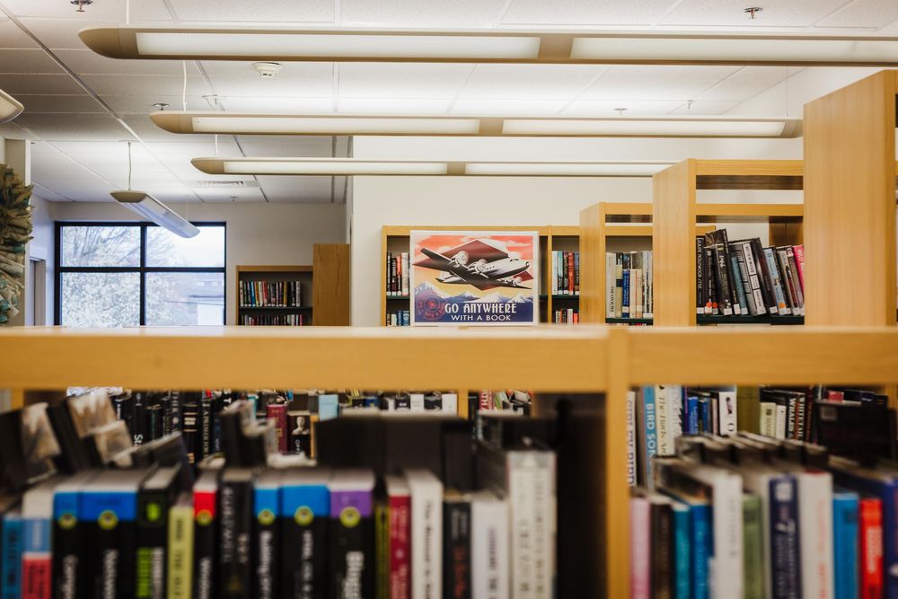 Library bookshelves with with "Go Anywhere With a Book" poster and windows visible through wooden partitions display rows of books.