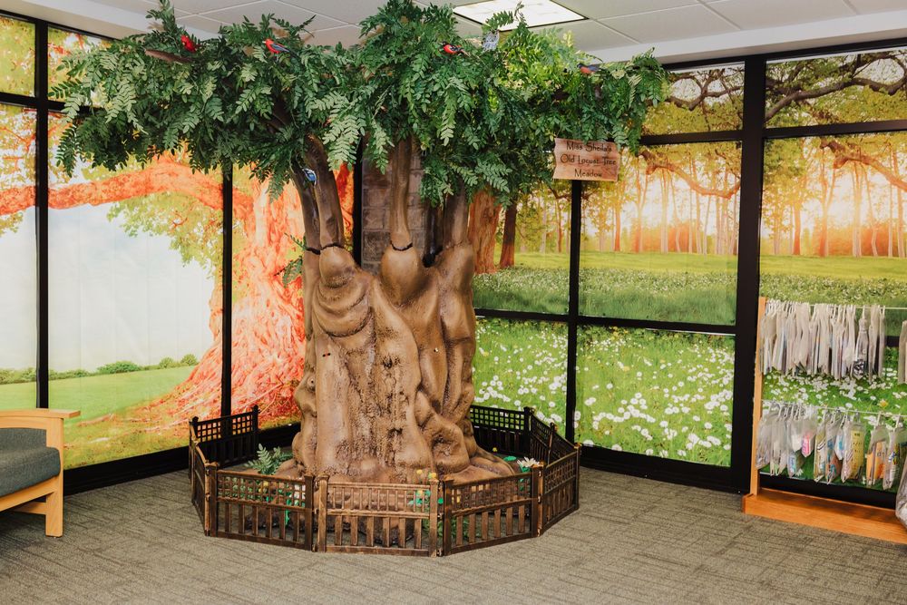 Large decorative indoor artificial tree with lush green foliage displayed near a rack of children's audio books at the library near me.
