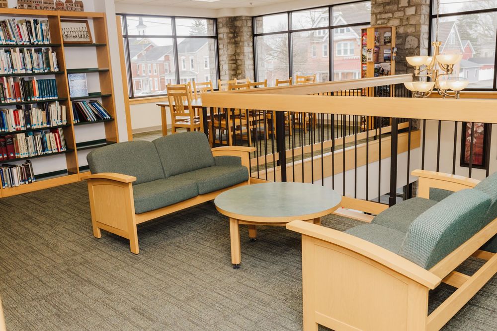 Library seating area with green cushioned benches and wooden coffee table surrounded by bookshelves and large windows with natural light.