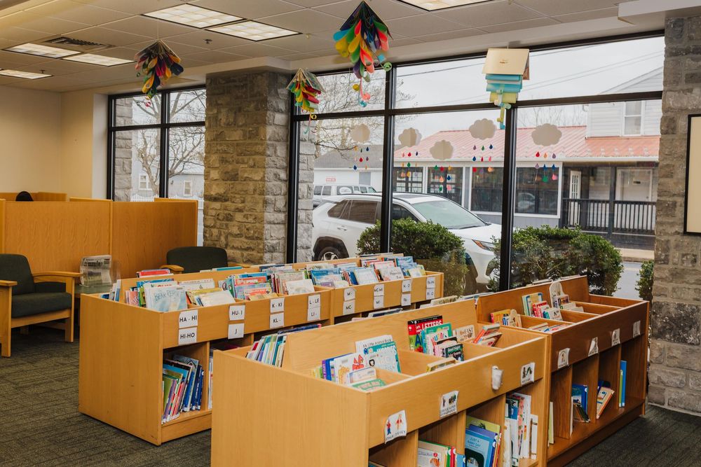 Wooden bookshelves with children's books line the windows of a bright library space decorated with colorful hanging decorations.