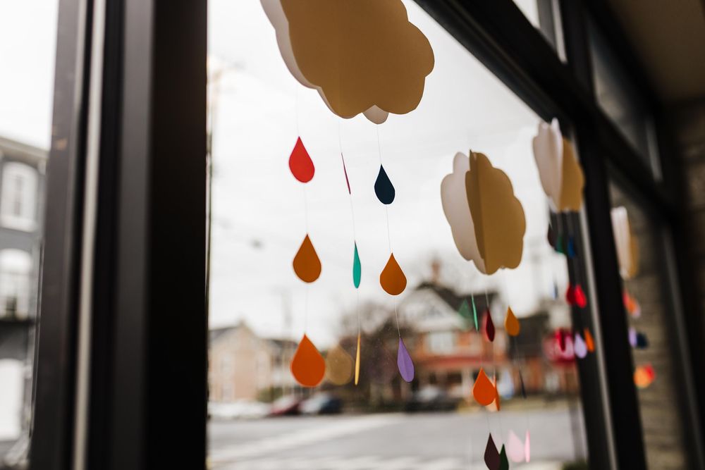 Colorful paper raindrops hang from a window frame overlooking a blurred city street outside.