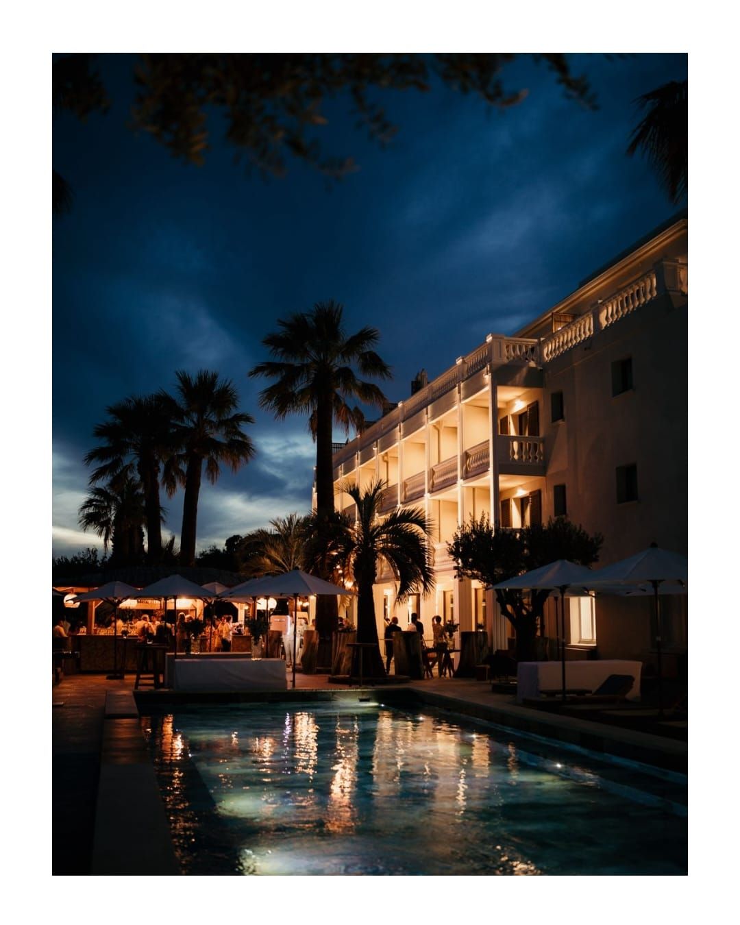 Illuminated resort hotel with palm trees and swimming pool reflecting in water during twilight hours at luxury destination.