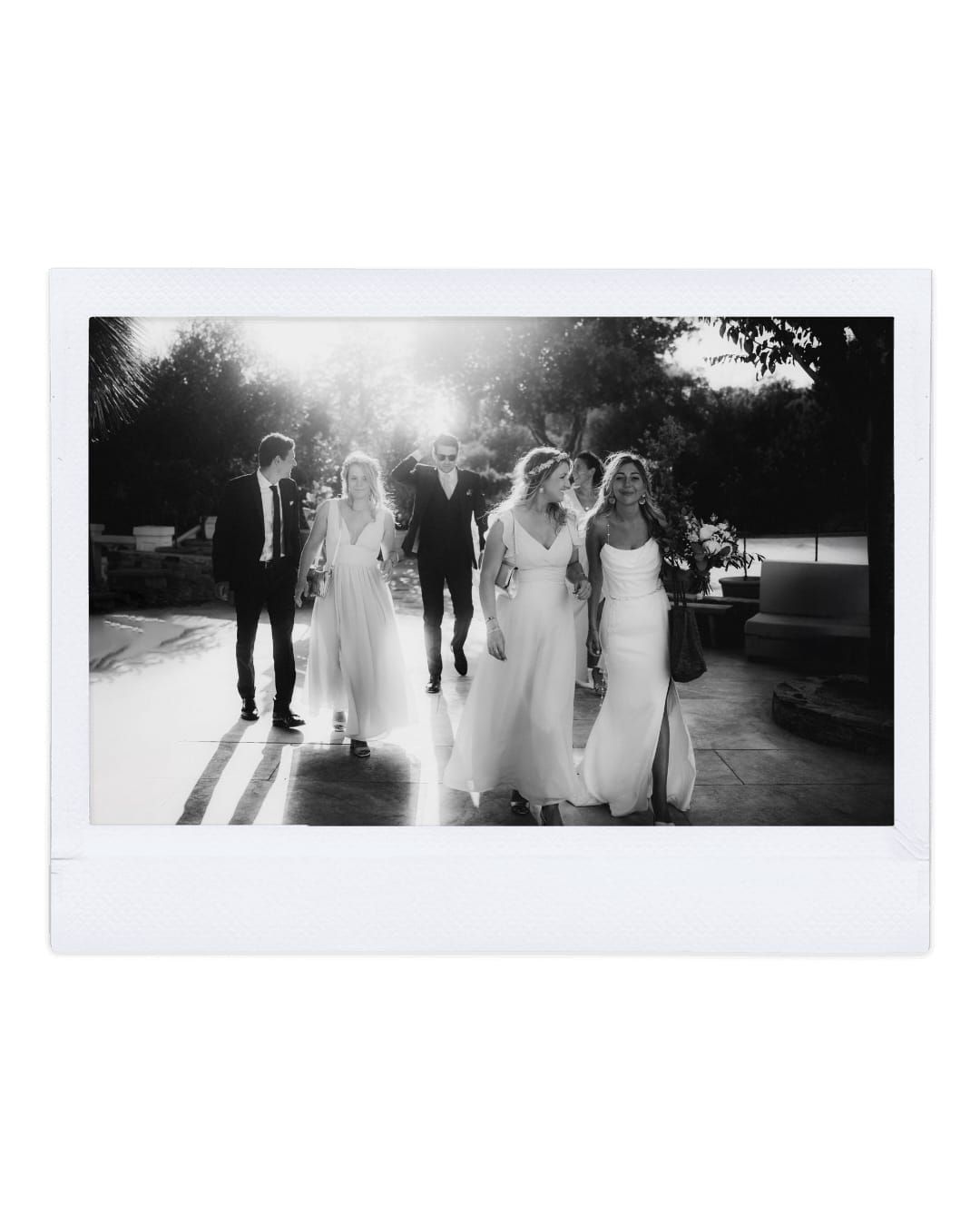 Black and white photo of wedding party walking together on sun-lit path outdoors.