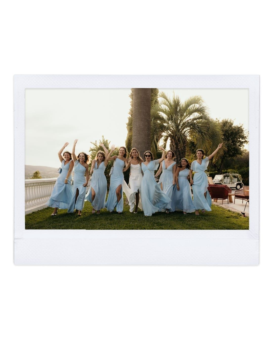A group in light blue bridesmaid dresses pose joyfully on a lawn with palm trees in the background.