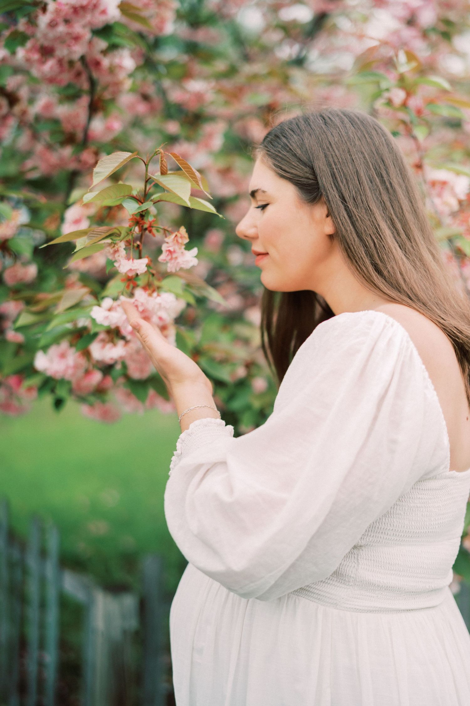 A woman in a flowing white dress admires delicate pink cherry blossoms in a spring garden.
