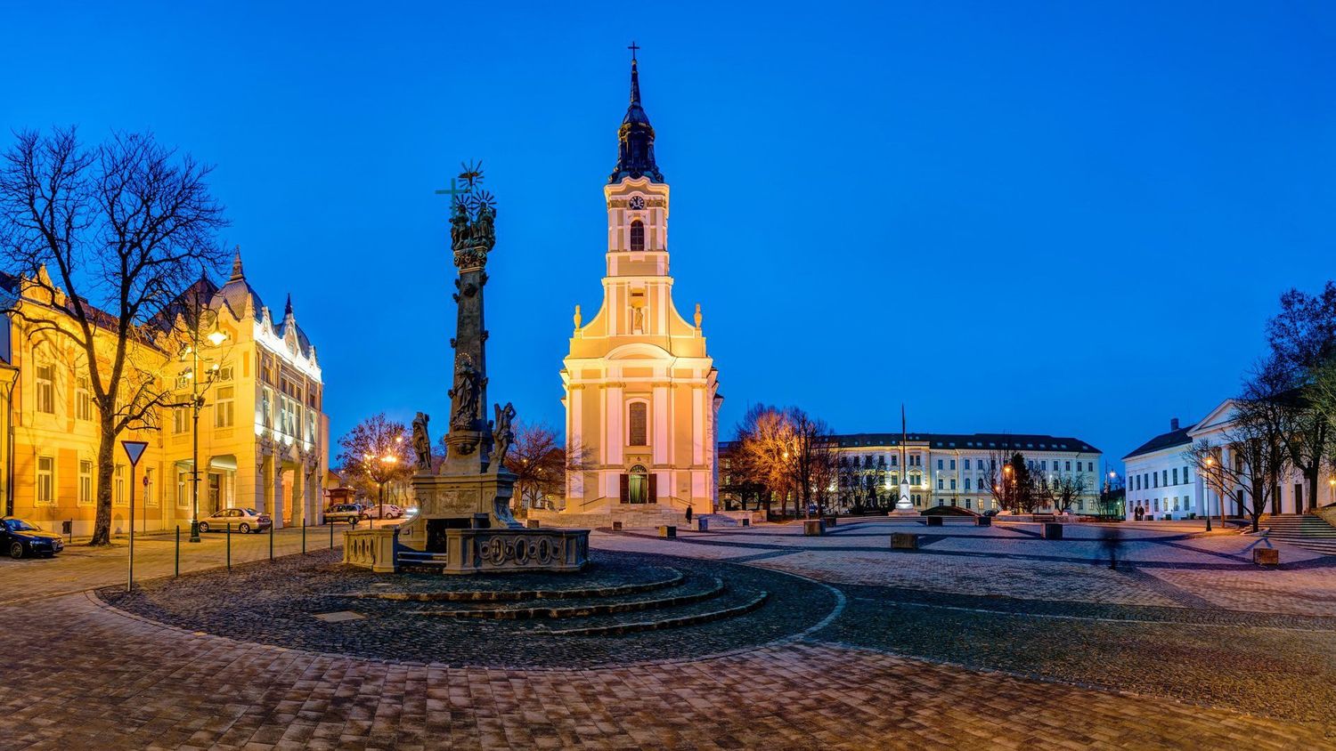 Illuminated town square in Zary, Poland with historic baroque church and plague column monument at dusk.