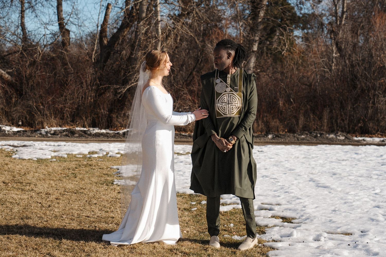 A couple stands together in winter scenery, with one in a white dress and one in traditional Scottish highland attire.