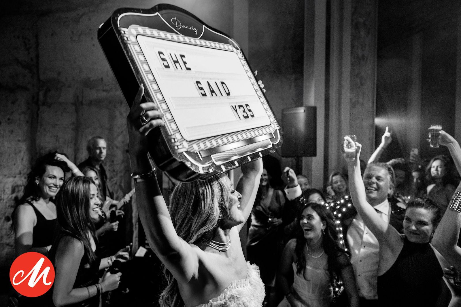 A black and white photo shows people gathered at a crowded bar, sharing an intimate moment amid bottles and glasses.