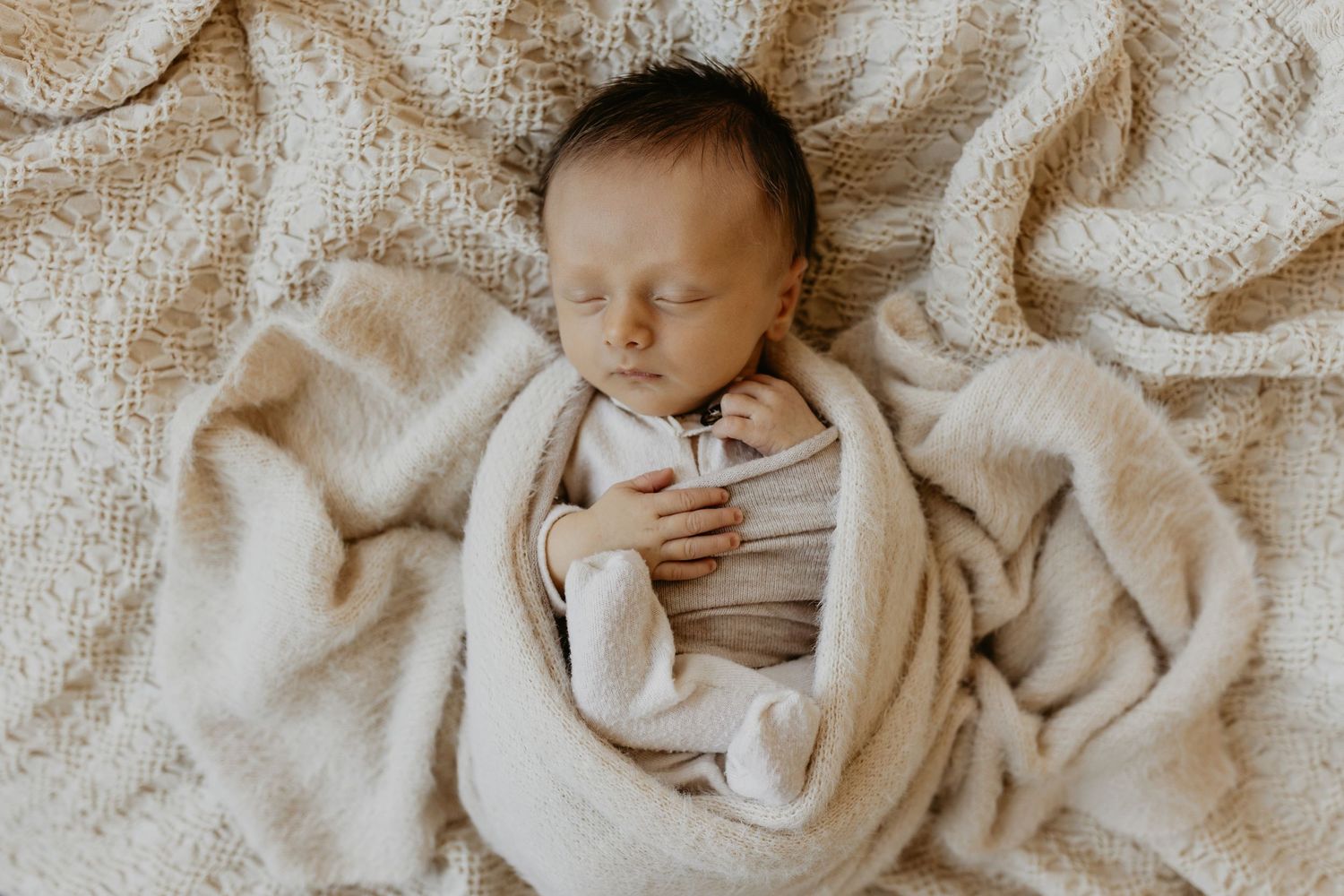 A sleeping newborn wrapped in soft beige swaddle blankets lies on textured cream bedding. Tulsa newborn photographer.
