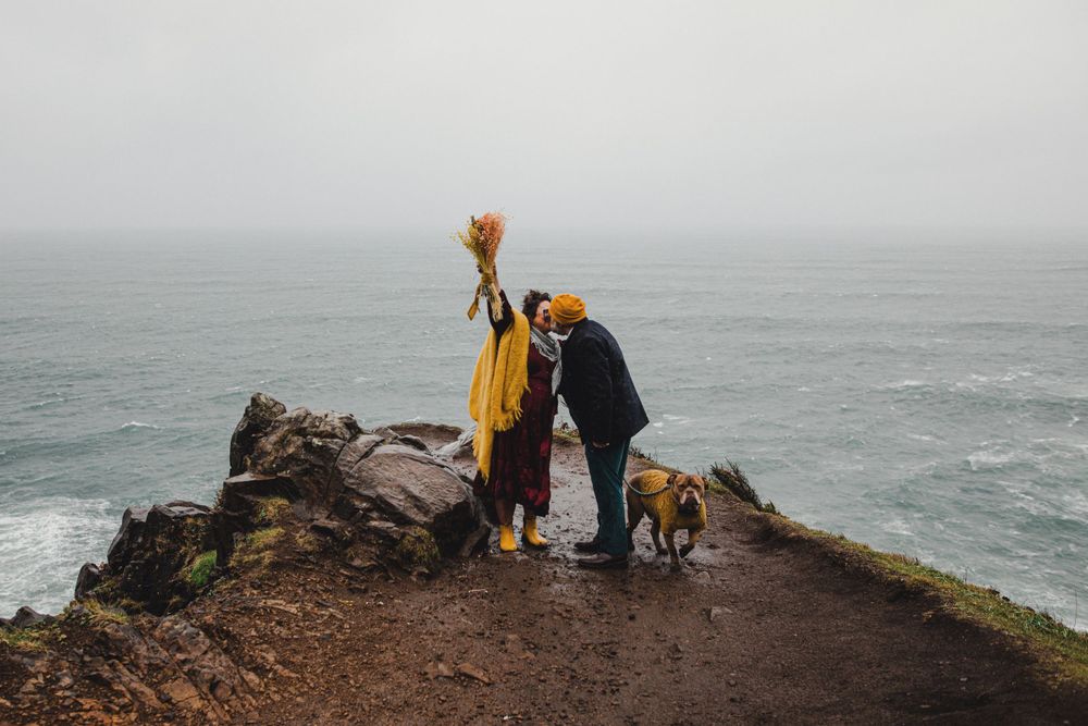 A couple and their dog stand at the edge of a dramatic coastal cliff overlook in yellow and black outfits.