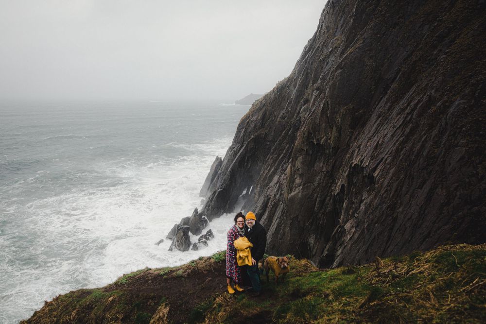 Person in yellow coat stands on steep coastal cliffs overlooking dramatic ocean waves.