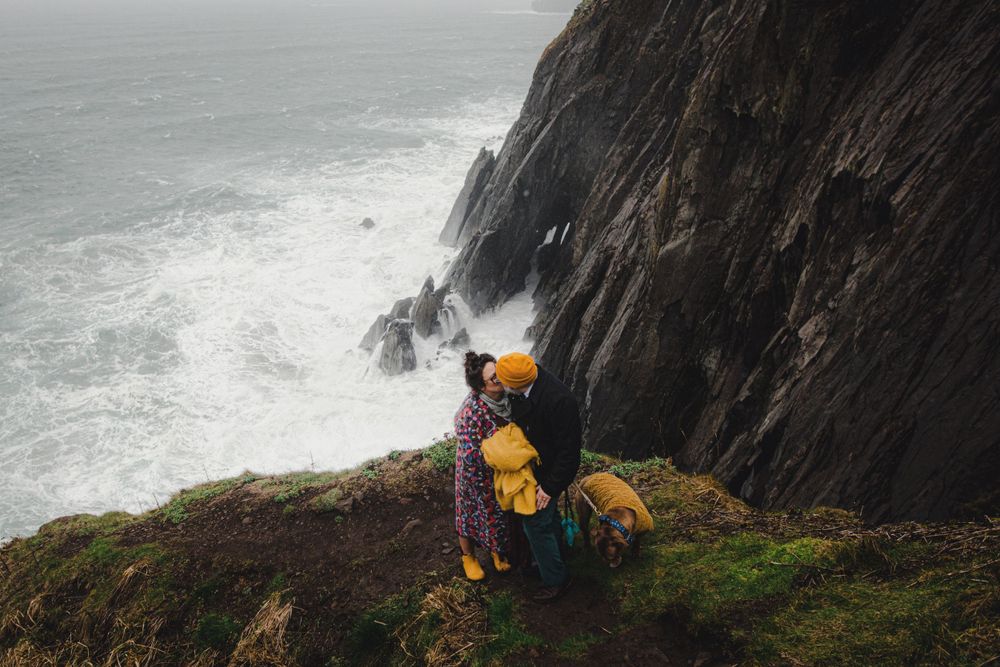 Person in yellow standing on steep cliff edge with dog watching waves crash below.