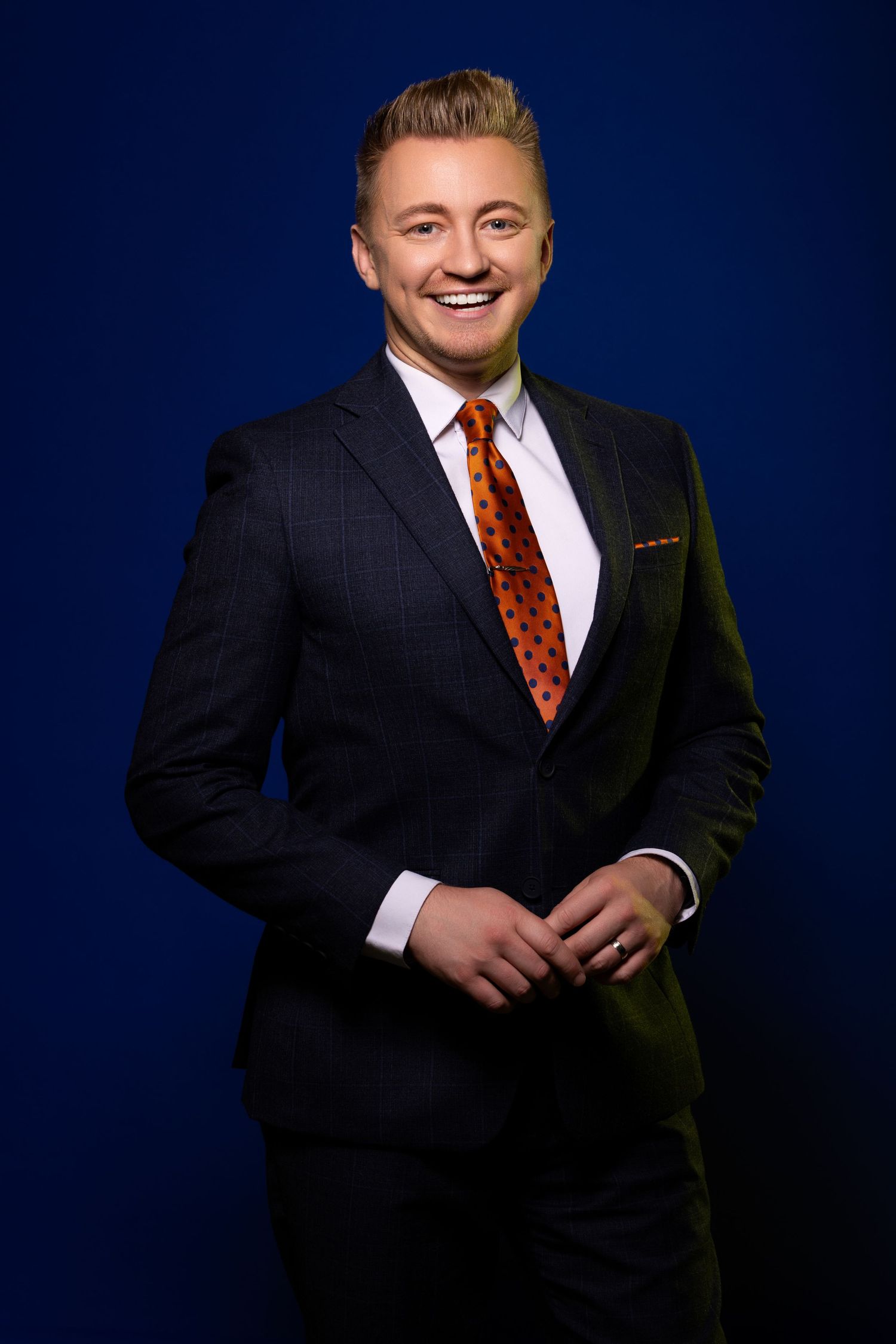 Professional studio portrait of person in dark suit with polka dot tie against blue background.