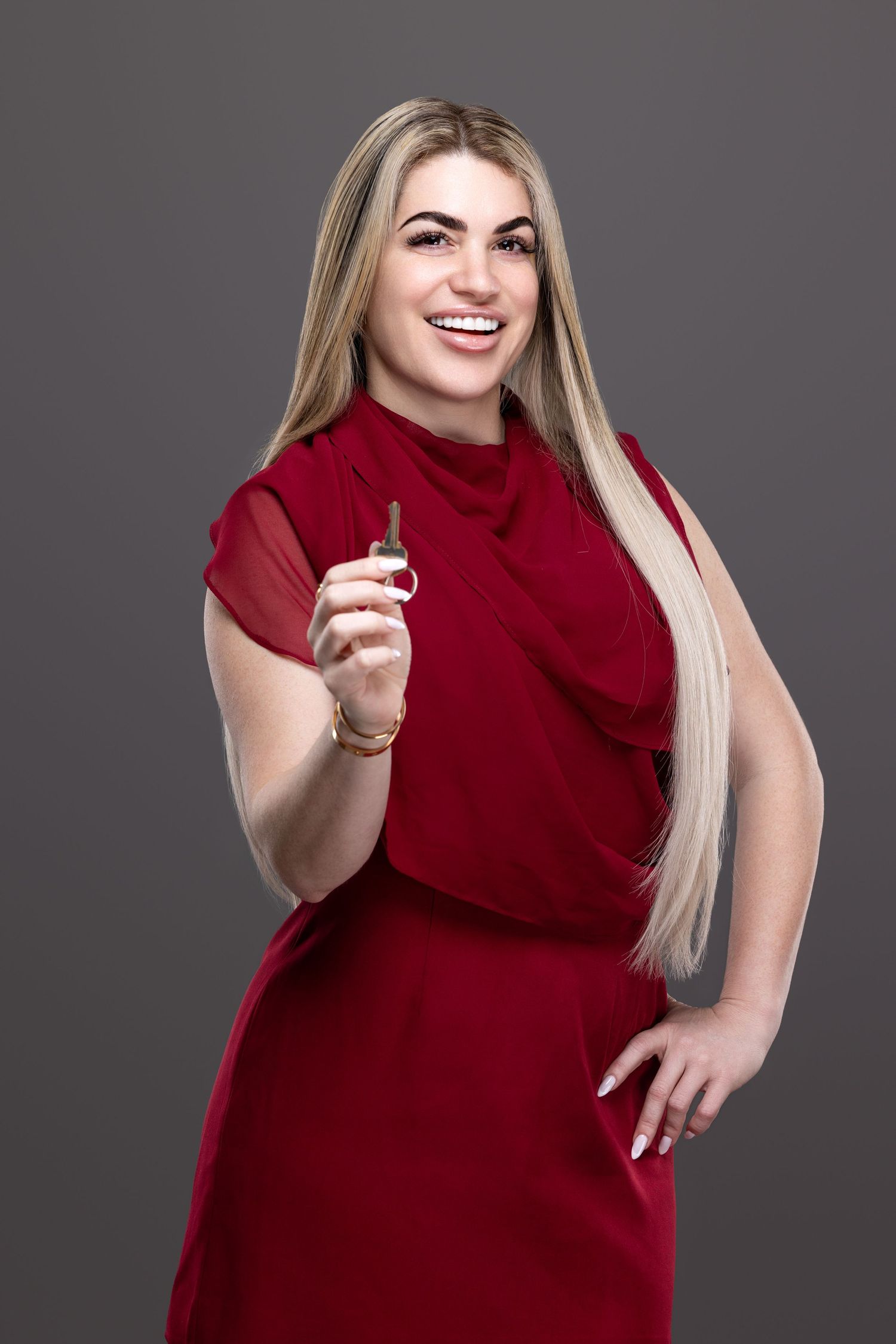 A person in a red dress poses confidently against a gray background while smiling and gesturing.