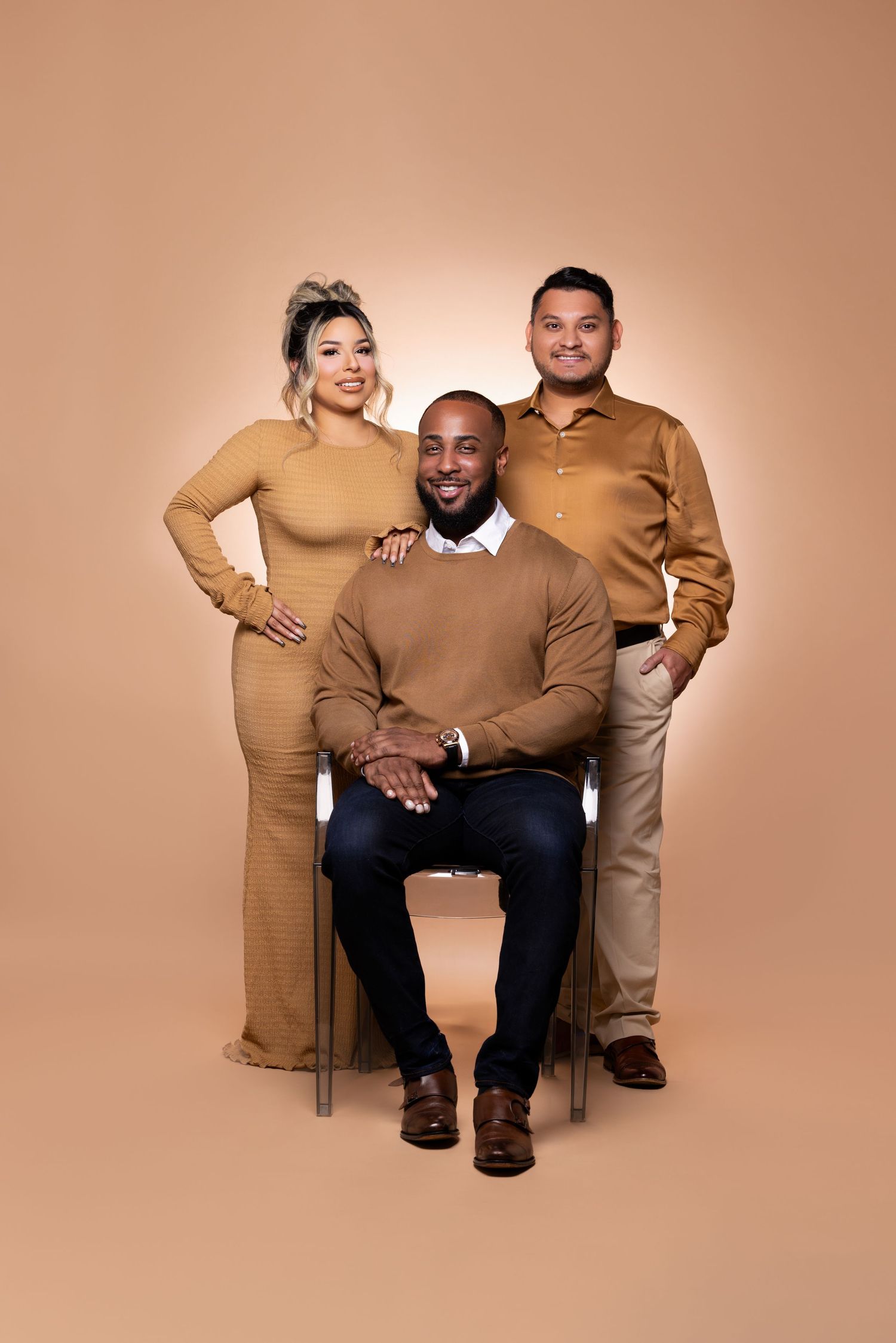 Three people in coordinated tan and brown outfits pose for a professional studio photo against a neutral backdrop.