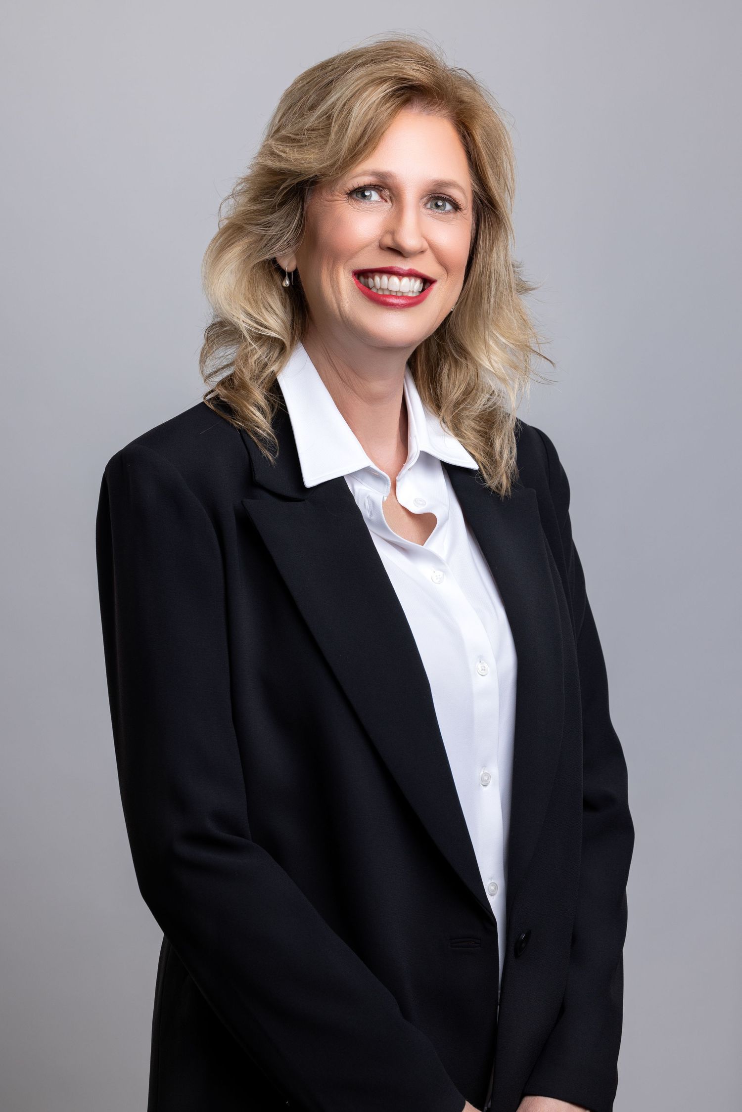Professional corporate headshot of person in black blazer and white blouse against gray background.