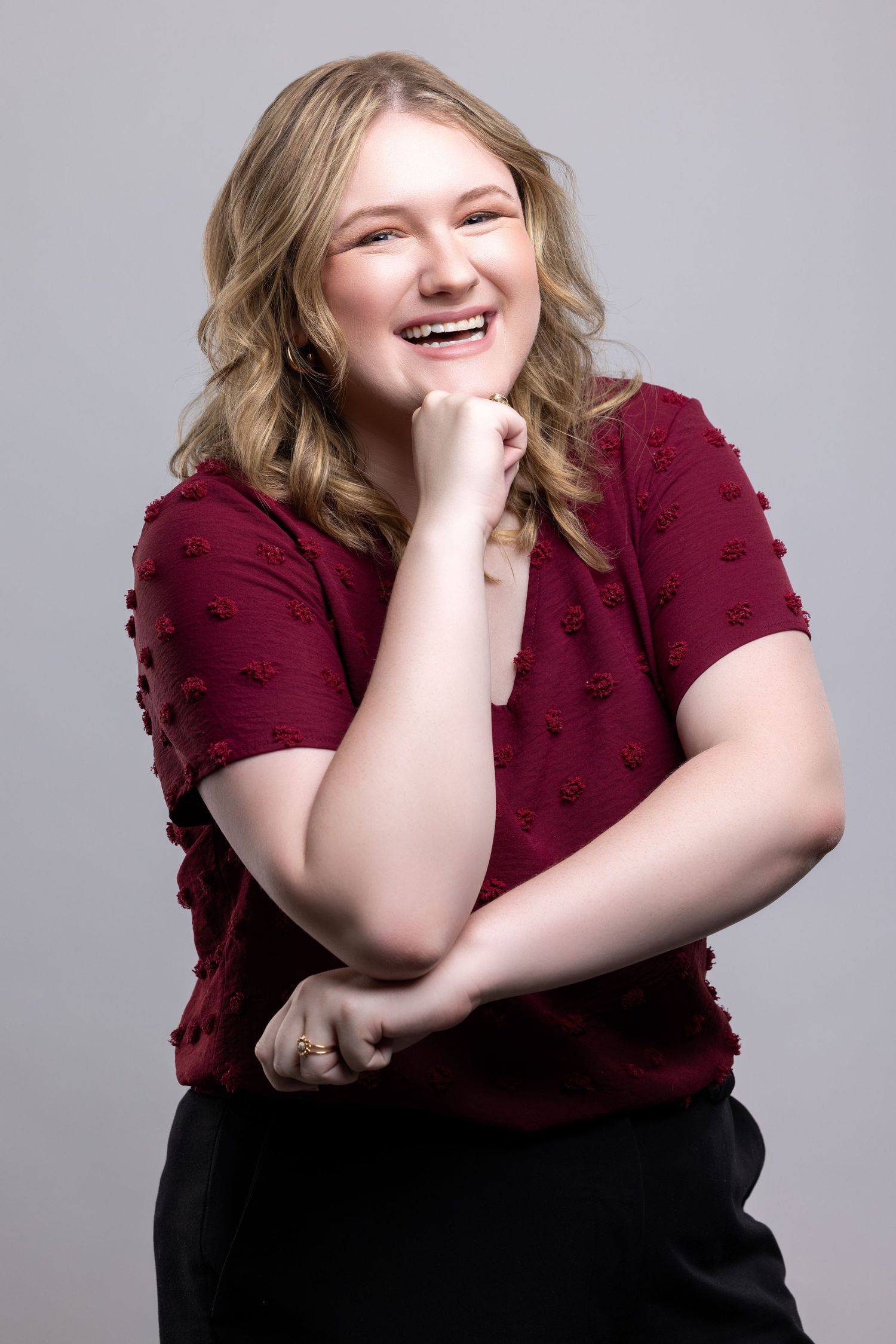 A joyful portrait of someone wearing a burgundy textured top posing against a gray background.