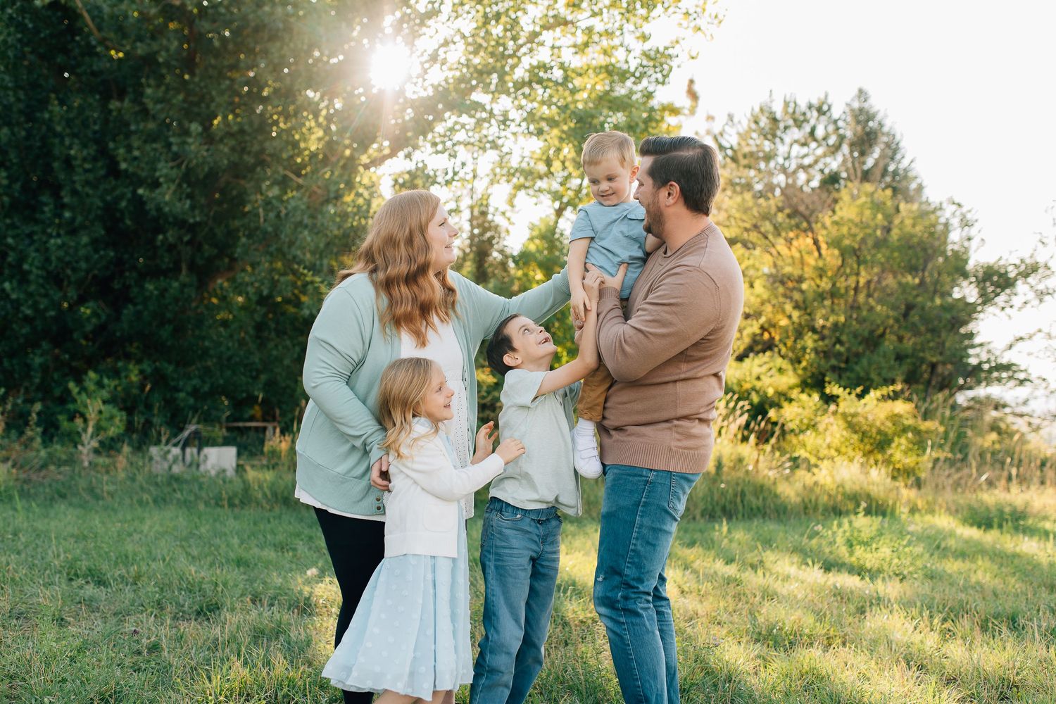 A family enjoys a tender moment together outdoors on a sunny evening in a grassy field surrounded by trees.