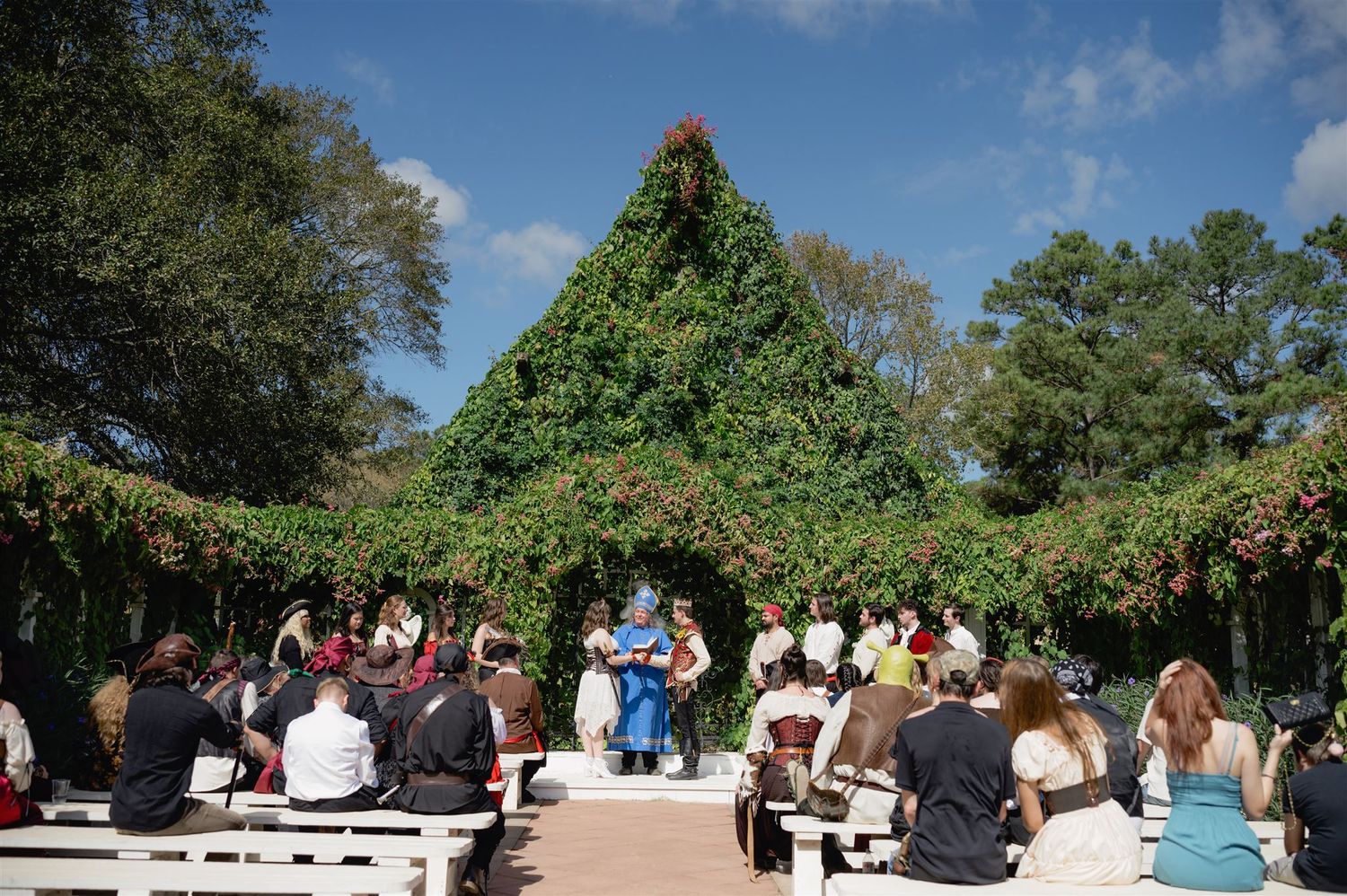 A panoramic view of an outdoor wedding ceremony with guests seated in rows under lush green trees and blue skies.
